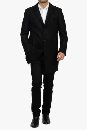 A man in a Howard Hudson Overcoat with black pants and a white dress shirt walks forward, one hand in his pocket, against a white background.