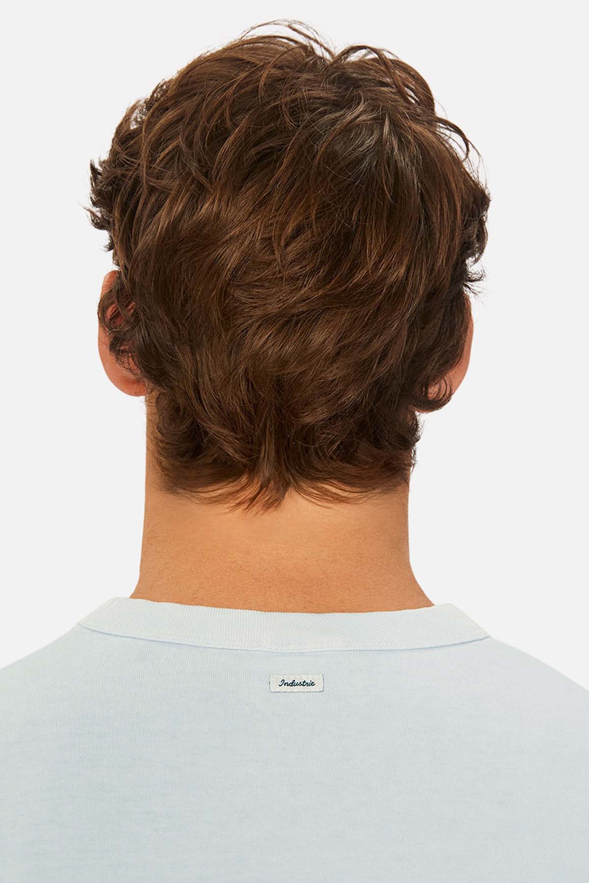 A person with wavy brown hair, seen from behind in a light blue Industrie The Del Sur Tee featuring a small white label on the collar, stands against a plain white background for use in size guides to help visualize fit and chest measurement.