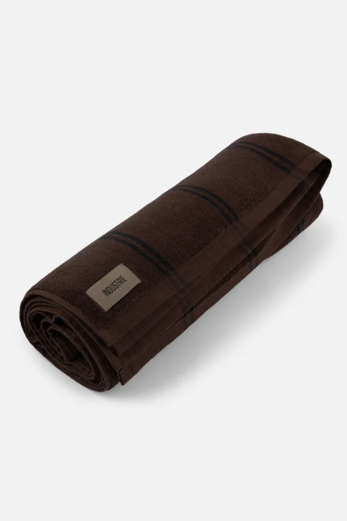 The Industrie The Comporta Beach Towel, made from soft cotton terry cloth in dark brown with a subtle plaid pattern and a rectangular stitched label, is shown rolled up on a plain light background.