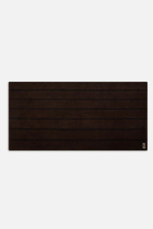 A rectangular, dark brown wooden surface with horizontal grooves and a small metal label in the bottom right corner sits against a plain white background, evoking the look of Industrie The Comporta Beach Towel’s cotton terry texture.