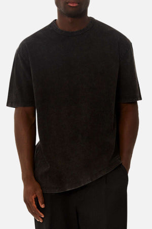 A man wears the Industrie The Del Sur Acid Wash Tee, an oversized, short-sleeved faded black t-shirt, paired with black pants, standing with one hand in his pocket against a plain white background.