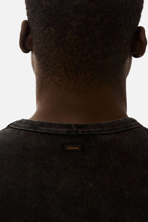 A close-up shows the back of a person with short hair wearing the Industrie The Del Sur Acid Wash Tee, an oversized faded black shirt with a small stitched neckline label, set against a plain light background.