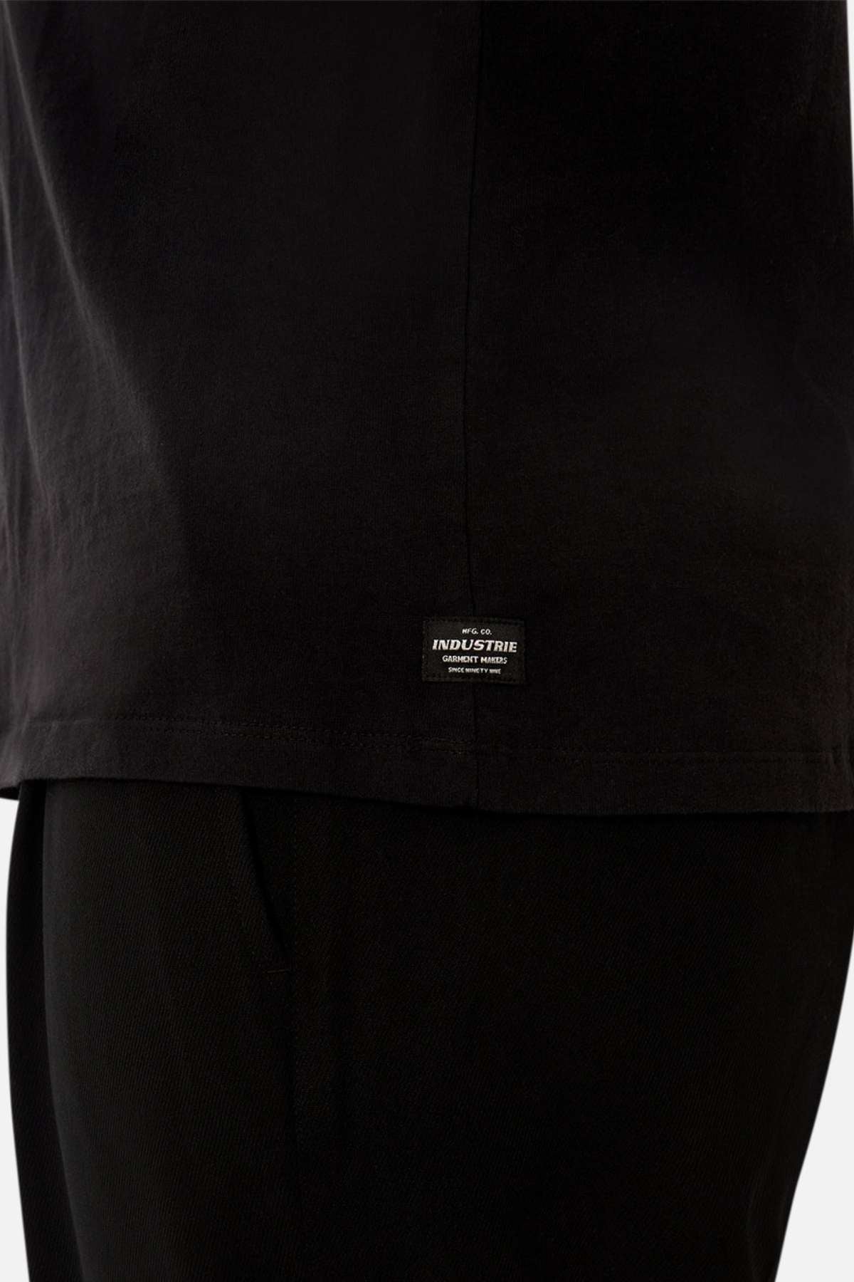 Close-up of a person wearing the Industrie The Del Sur Sleeveless Tee in black, featuring an oversized fit and a small rectangular "Industrie" label near the hem. Paired with black pants against a plain white background.