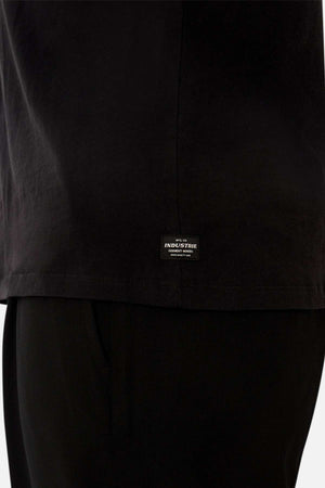 Close-up of a person wearing the Industrie The Del Sur Sleeveless Tee in black, featuring an oversized fit and a small rectangular "Industrie" label near the hem. Paired with black pants against a plain white background.