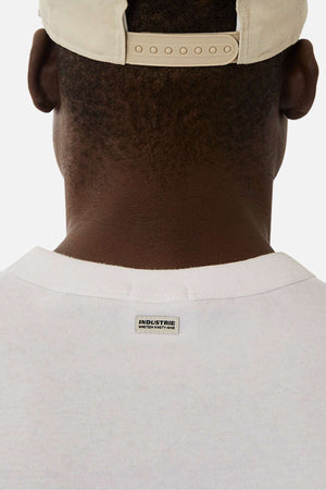 A close-up of the back of a person wearing the Industrie The Del Sur Sleeveless Tee in white, made from 100% cotton with an "INDUSTRIE" label on the collar and a beige baseball cap, set against a plain white background.