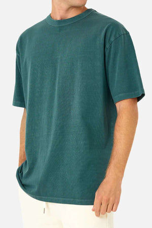 Wearing the Industrie The Del Sur Tee—an oversized, 100% cotton teal shirt—paired with off-white pants, a person stands against a plain light background, one hand in their pocket and their head partially out of frame.