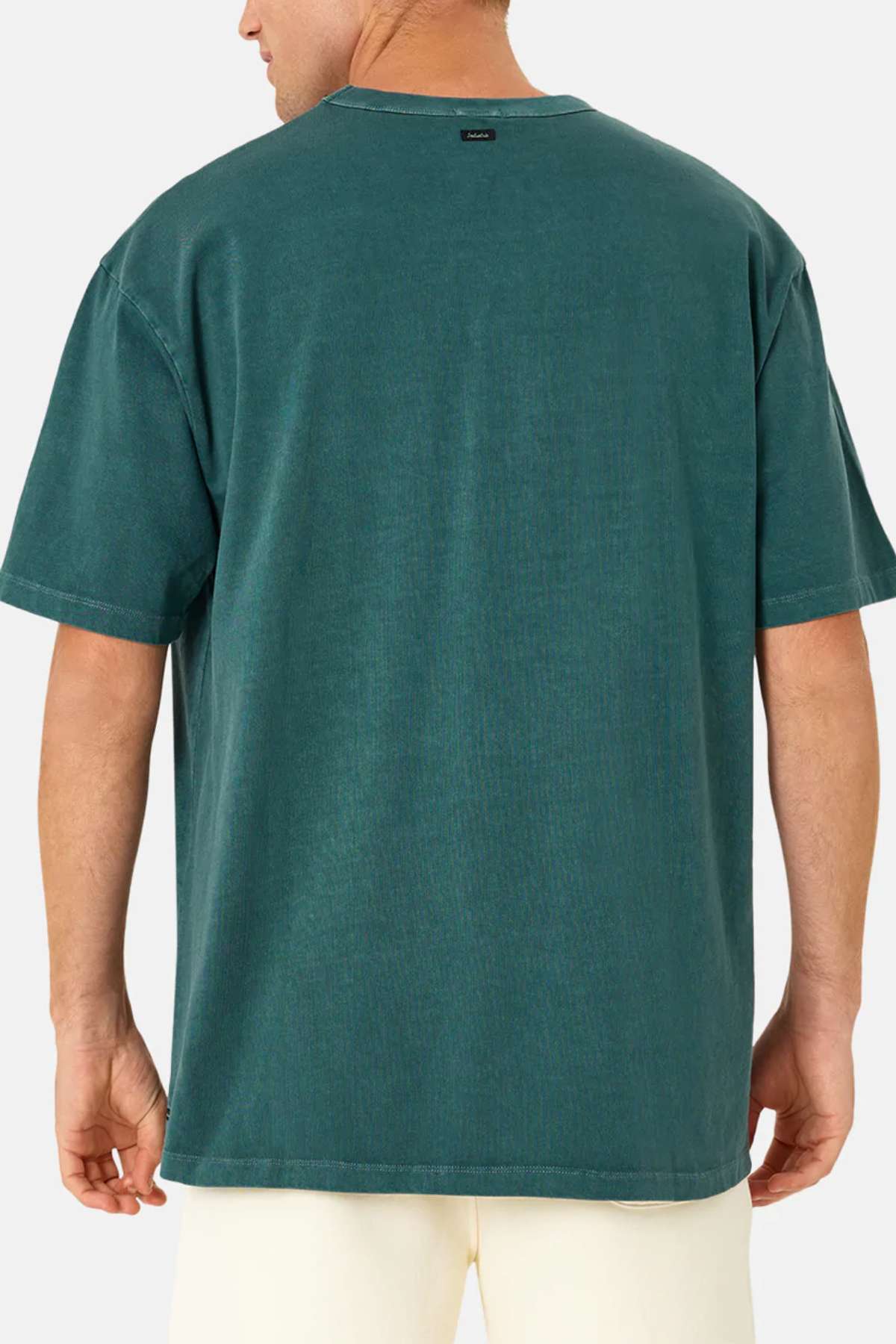 A man is seen from behind wearing the Industrie The Del Sur Tee, an oversized short-sleeve teal shirt made of 100% cotton, paired with light-colored shorts against a plain background.