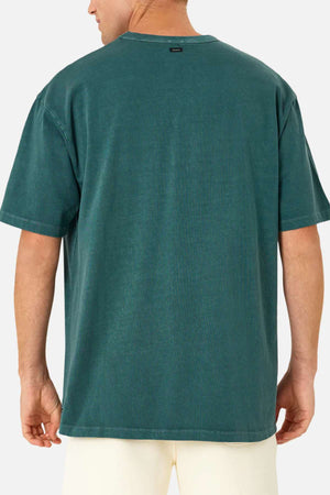 A man is seen from behind wearing the Industrie The Del Sur Tee, an oversized short-sleeve teal shirt made of 100% cotton, paired with light-colored shorts against a plain background.
