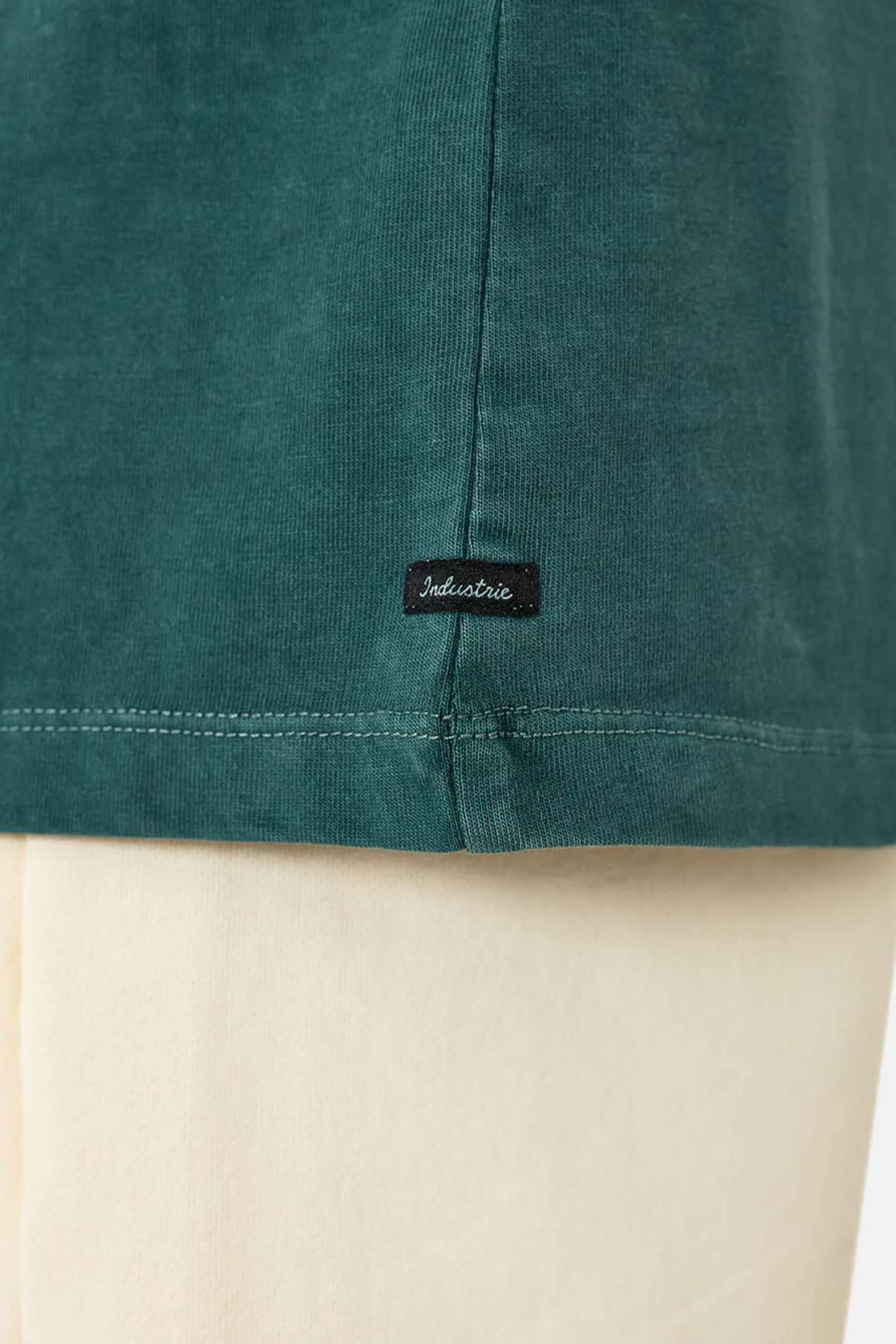 Close-up of the bottom hem of the Industrie The Del Sur Tee in green, featuring a small black "Industrie" tag. Worn over cream-colored pants and crafted from 100% cotton for comfort and style.