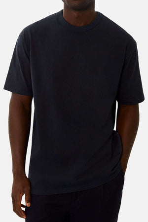 A person models the Industrie The Del Sur Tee—an oversized acid wash shirt—paired with dark pants, standing against a light background with one hand in their pocket. Their face is partially obscured.