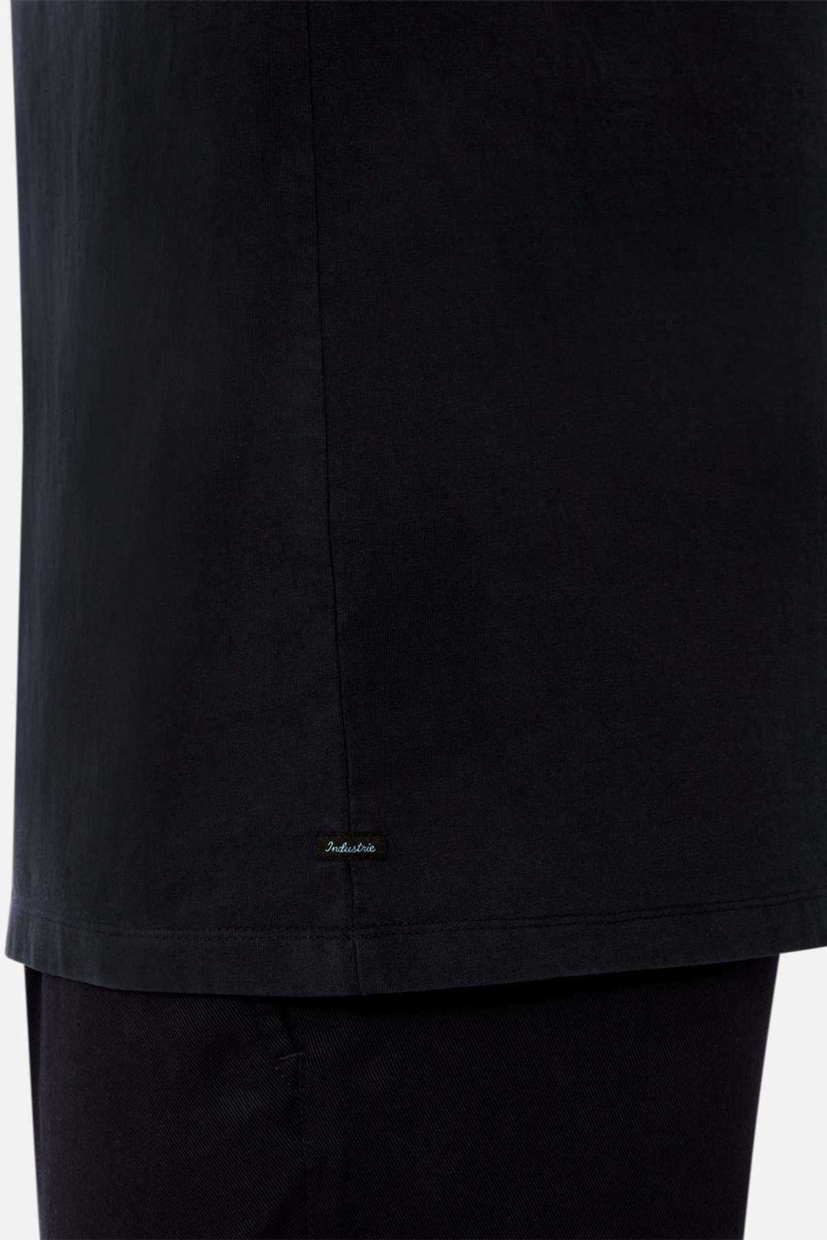 Close-up of a person wearing the Industrie The Del Sur Tee in black, an oversized 100% cotton shirt with a small embroidered label near the hem, paired with black pants. The background is plain and light-colored.