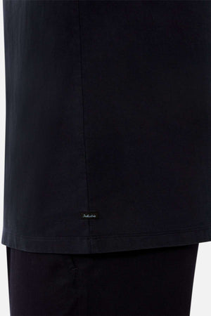 Close-up of a person wearing the Industrie The Del Sur Tee in black, an oversized 100% cotton shirt with a small embroidered label near the hem, paired with black pants. The background is plain and light-colored.