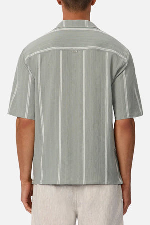 A person wearing the Industrie The Fullerton Short Sleeve Shirt—a light gray, collared men's shirt with vertical white stripes—seen from the back, paired with light-colored shorts for a versatile look.