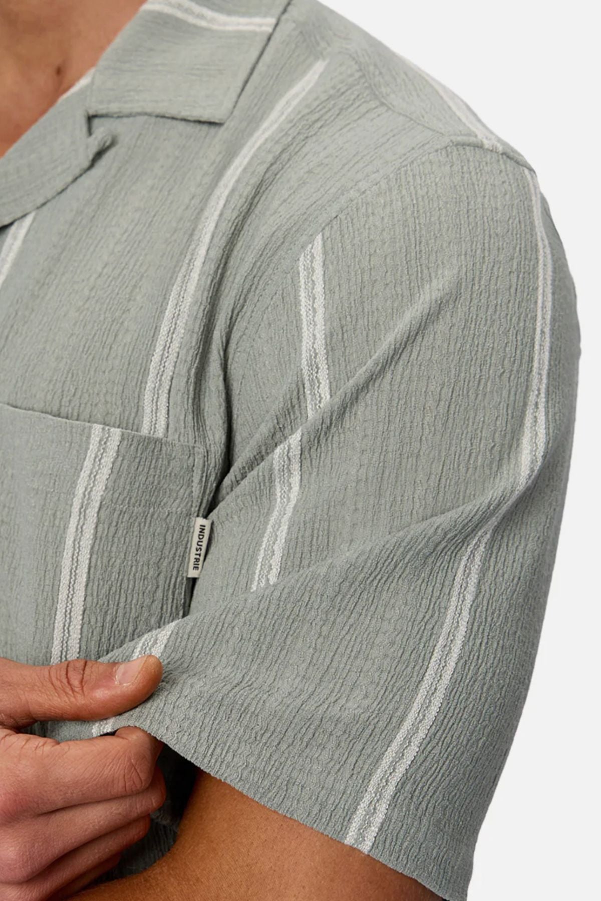 A person wears the Industrie The Fullerton Short Sleeve Shirt in light green with white vertical stripes, short sleeves, a pocket, and a small fabric brand tag—making it a versatile men's wardrobe essential.