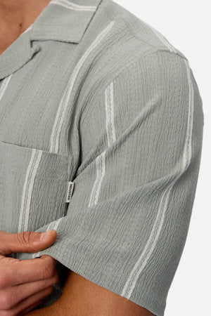 A person wears the Industrie The Fullerton Short Sleeve Shirt in light green with white vertical stripes, short sleeves, a pocket, and a small fabric brand tag—making it a versatile men's wardrobe essential.