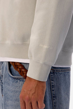 Someone is wearing the Industrie The New Del Sur Washed Sweat in an oversized fit with subtle wrist embroidery, paired with blue jeans, a brown braided belt, and a white shirt peeking out. Only their torso and arm are visible.