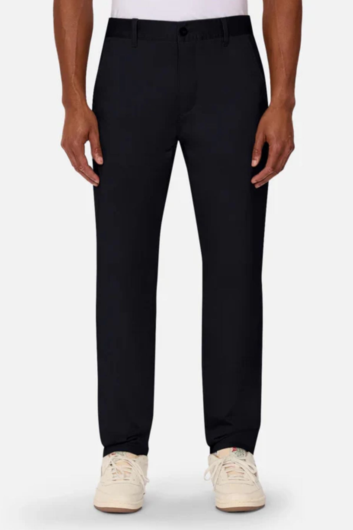 A person wears Industrie The Regular Cuba Chino Pant in black, paired with a white shirt and white sneakers, standing against a plain light background. The photo captures the look from shoulders to feet with arms relaxed at their sides.