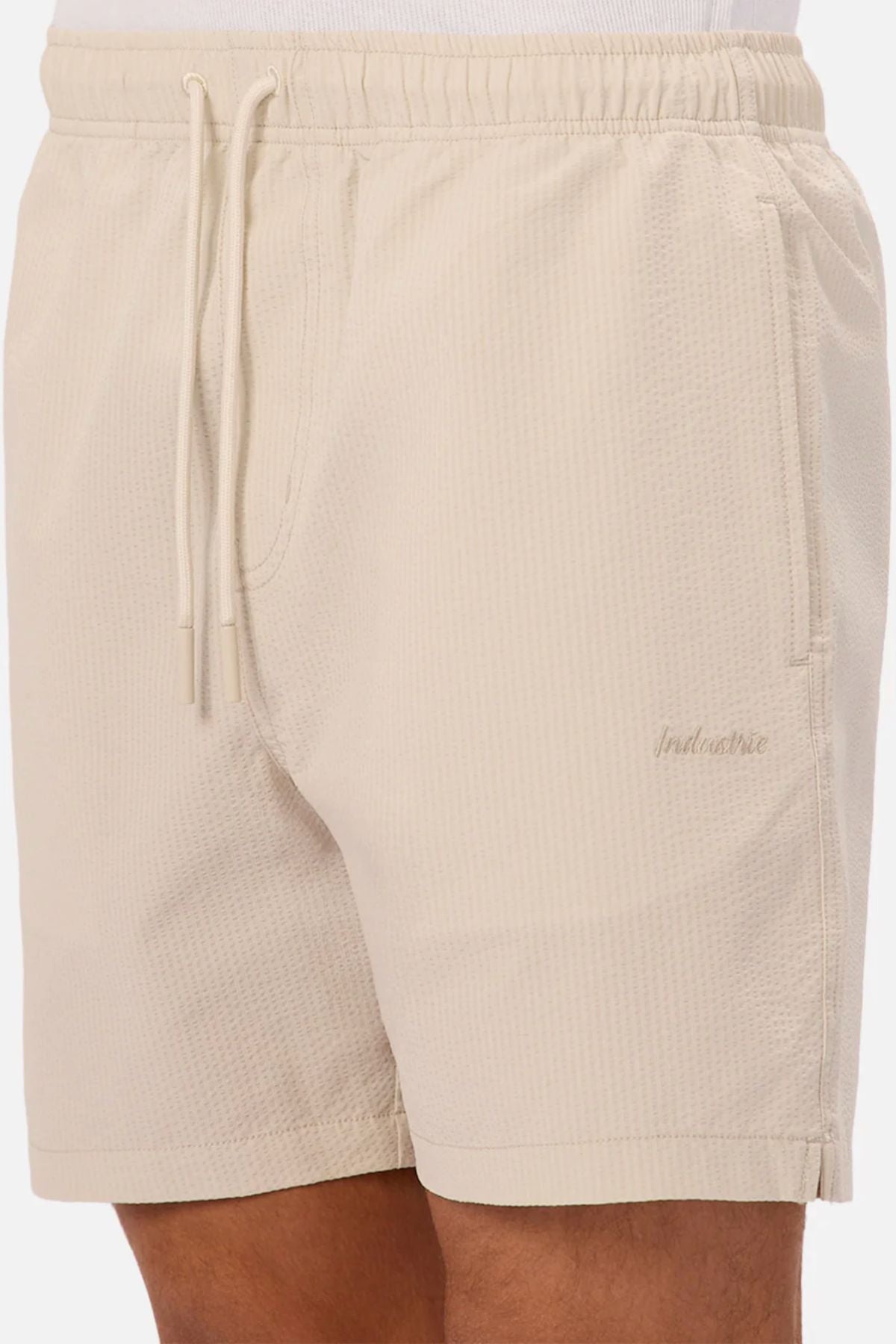 A person wears Industrie The New Seersucker Swim Short in light beige with textured quick-dry polyester, elastic waistband, drawstring, side pocket, and subtle "Industrie" logo on the left thigh. Only lower torso and upper legs are visible.