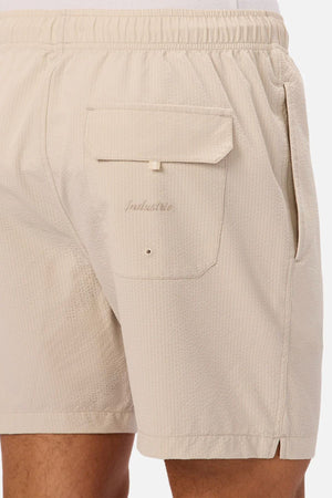 A close-up view of a person wearing Industrie The New Seersucker Swim Short in light beige, highlighting the back pocket with flap and button detail alongside a side pocket.
