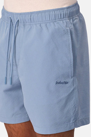 A close-up of a person in Industrie The New Seersucker Swim Short—light blue with textured lines and "Industrie" embroidery. Made from quick-dry polyester, these men's swim shorts are ideal for summer.