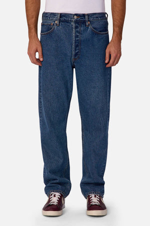 A person wearing Industrie The 90's Jean in vintage washed denim, a tucked-in white T-shirt, and maroon sneakers with white laces stands against a plain white background.