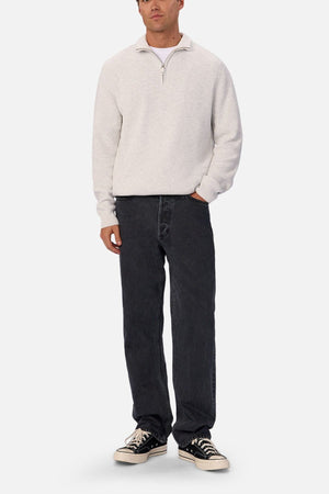 A person stands against a plain background wearing a light gray quarter-zip sweater, Industrie The 90's Jean in vintage-washed denim, and black low-top sneakers with white soles.