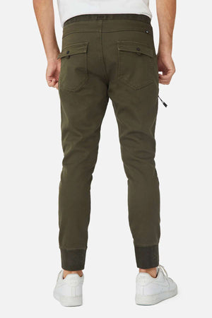 A person is seen from behind wearing olive green Industrie The Armoured Drifter Pant joggers with buttoned back pockets and ribbed cuffs, paired with white sneakers—ideal for the modern adventurer.