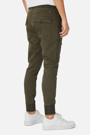 A person is seen from the back and side wearing Industrie The Armoured Drifter Pant—olive green cargo joggers with cuffed ankles and pockets—paired with white sneakers, against a plain white background, perfect for modern adventurers.