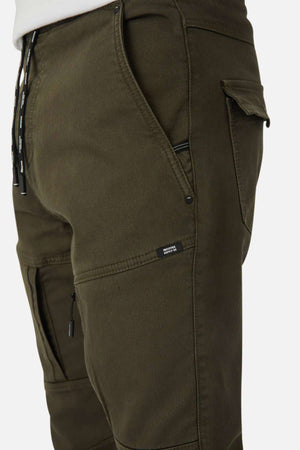 Side view of a person wearing Industrie The Armoured Drifter Pant in olive green, featuring roomy cargo and zippered pockets, a drawstring waist, and a "BRAVE SOUL" tag—ideal for any adventurer.