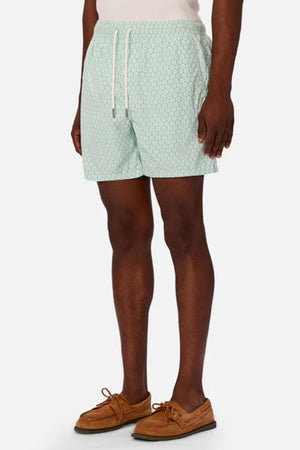 A man wearing Industrie The Aruba Swim Short in light green pattern, paired with a white shirt and brown boat shoes, stands with his hands by his sides against a plain white background.