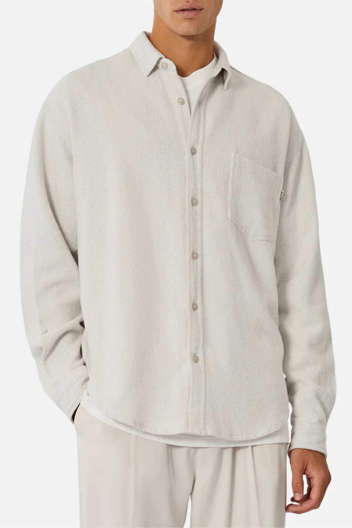 A man wears the Industrie The Atacama Shirt—a light beige 100% cotton button-up with a chest pocket—over a white tee and matching pants, standing with one hand in his pocket for a classic style.