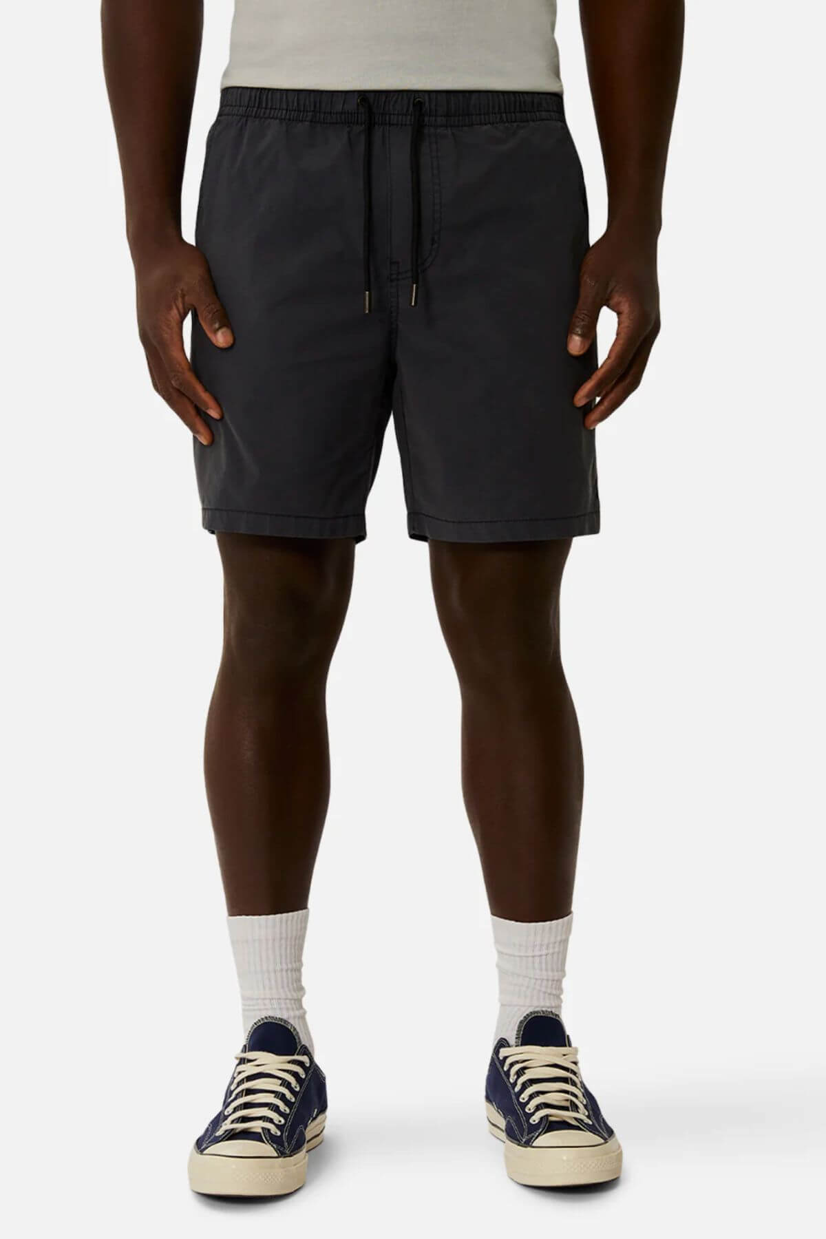 A person stands against a plain background wearing a light gray shirt, white crew socks, navy blue sneakers with white laces, and dark gray Industrie The Bahama Angler Shorts with an elastic waist.