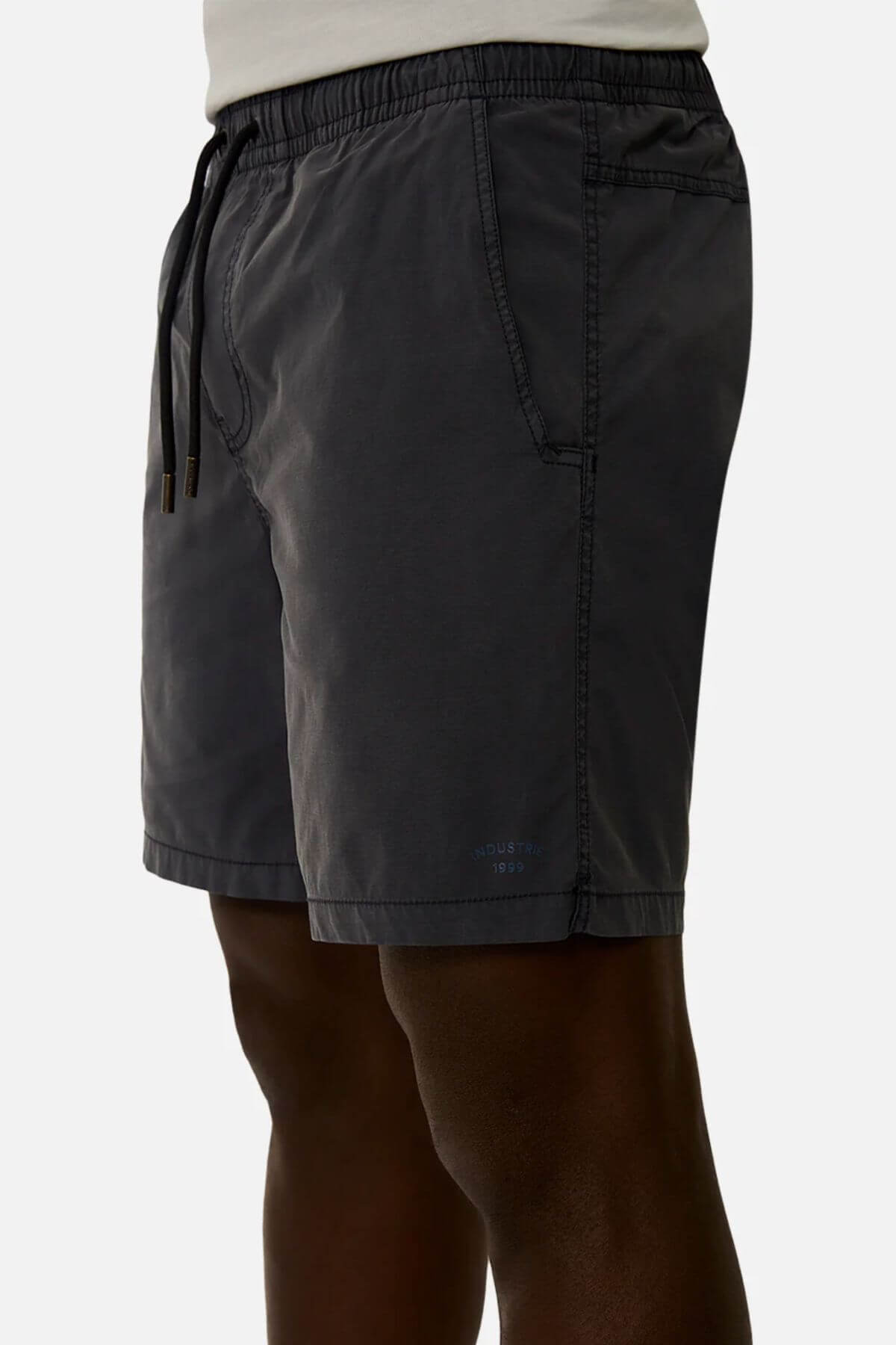 A person wears Industrie The Bahama Angler Short in dark gray, featuring an elastic waist, side pockets, and the Industrie logo, standing against a plain light background.