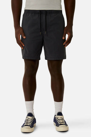 A person stands against a plain background wearing a light gray shirt, white crew socks, navy blue sneakers with white laces, and dark gray Industrie The Bahama Angler Shorts with an elastic waist.