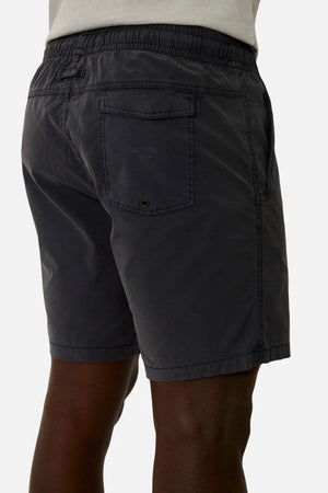 Someone stands with their back to the camera, wearing Industrie The Bahama Angler Short—dark gray swim shorts with an elastic waist, a back pocket, and the Industrie logo—against a plain backdrop.