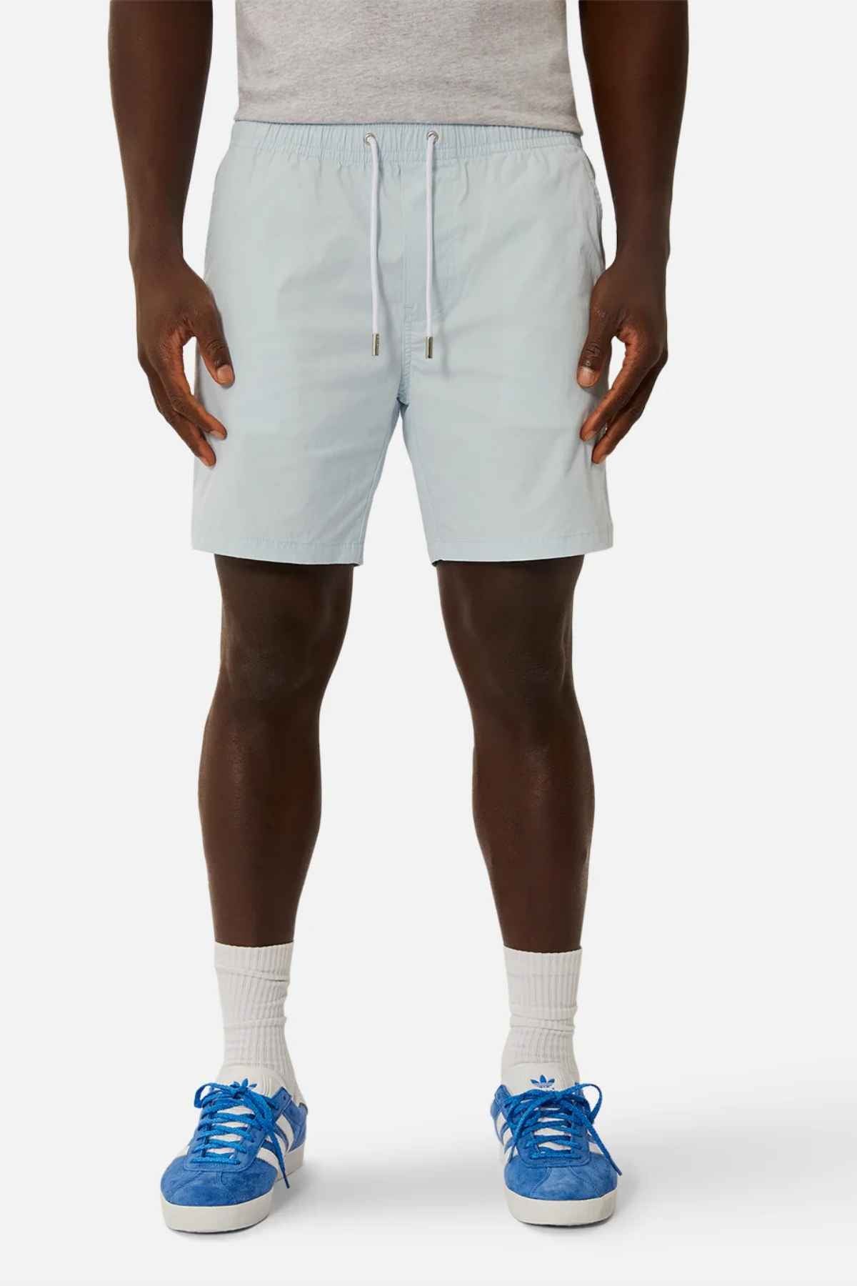 A person wearing light blue Industrie The Bahama Angler Short with a drawcord waist, a gray shirt, white socks, and blue sneakers stands against a plain white background.