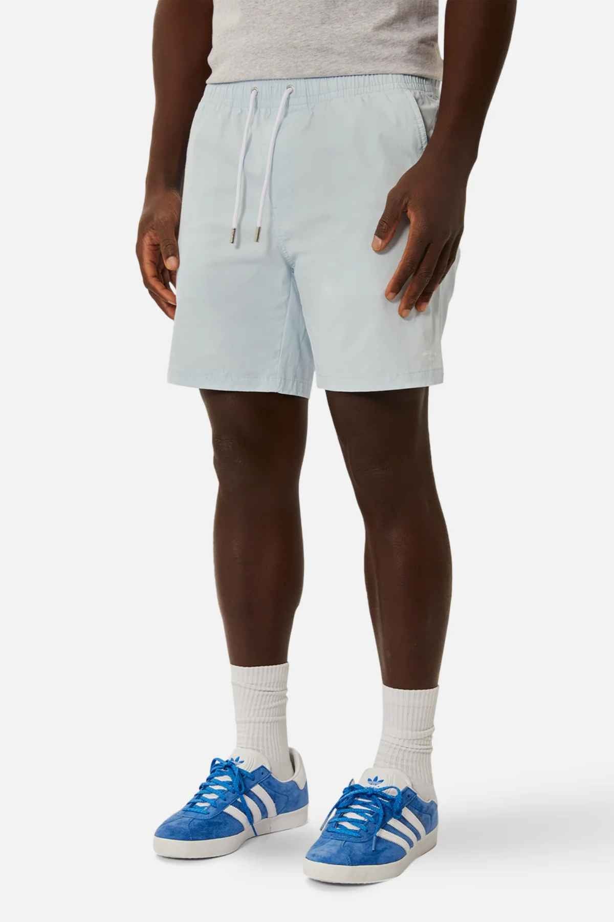 A person wearing a gray t-shirt and Industrie The Bahama Angler Short, which features a drawcord waist, complemented by white socks and blue Adidas sneakers, stands against a plain white background.