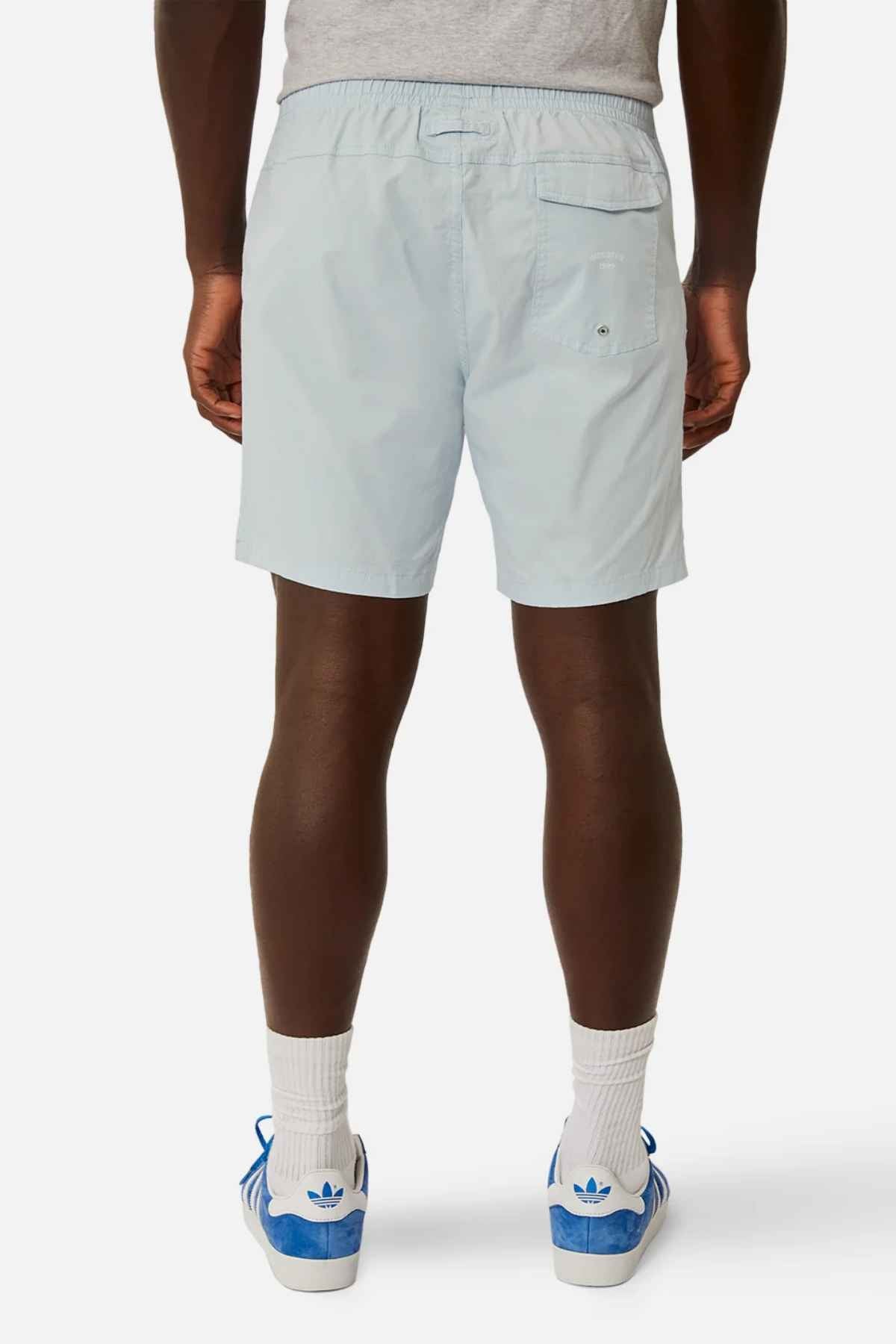A person stands facing away, wearing light blue Industrie The Bahama Angler Shorts with a drawcord waist and a back pocket with a button, paired with a gray shirt, white socks, and blue Adidas sneakers. The background is plain white.