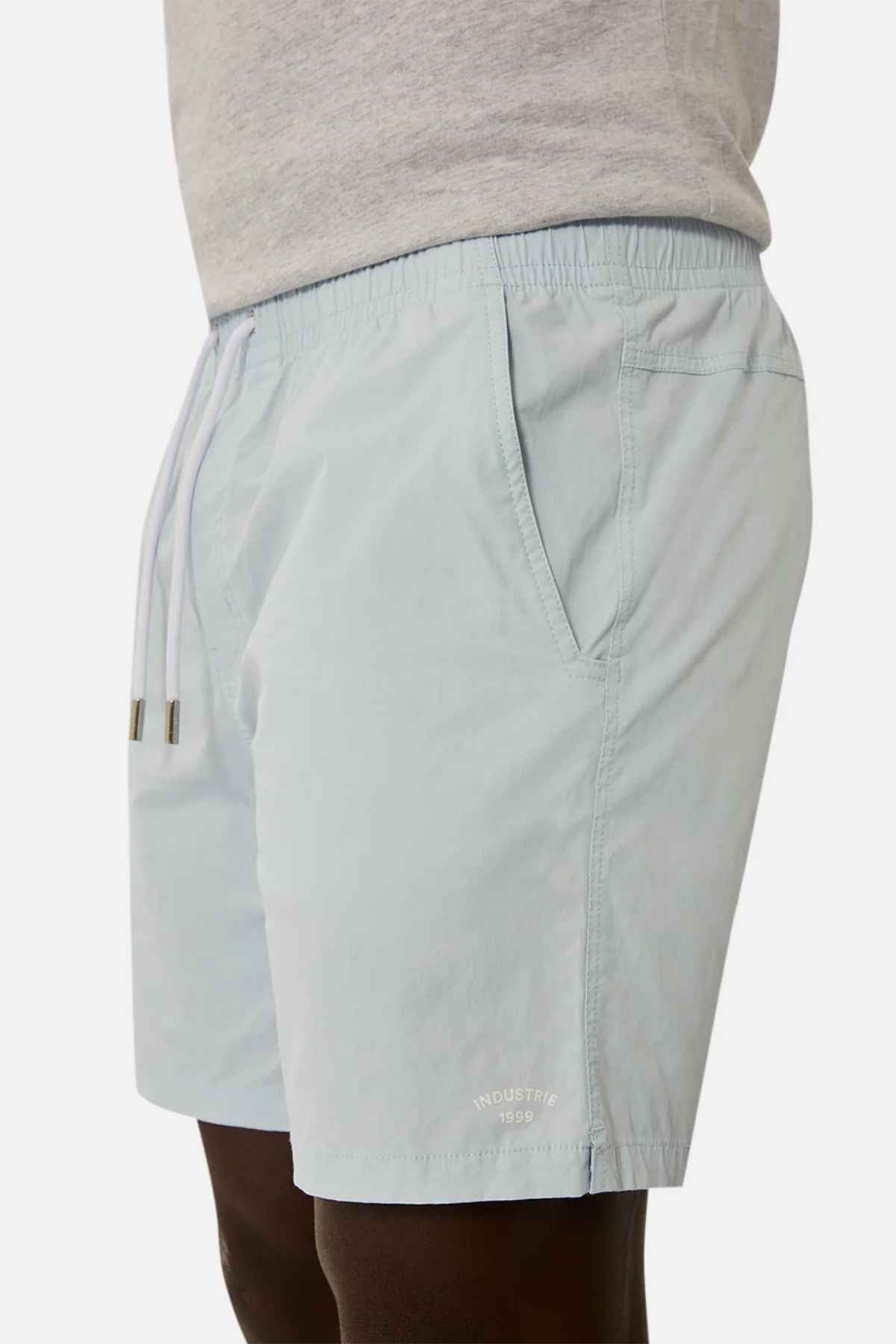A person is seen in light blue Industrie The Bahama Angler Shorts, complete with an elastic drawcord waist and side pockets, showcasing the "Industrie 1999" branding near the hem. They complement their outfit with a gray shirt.
