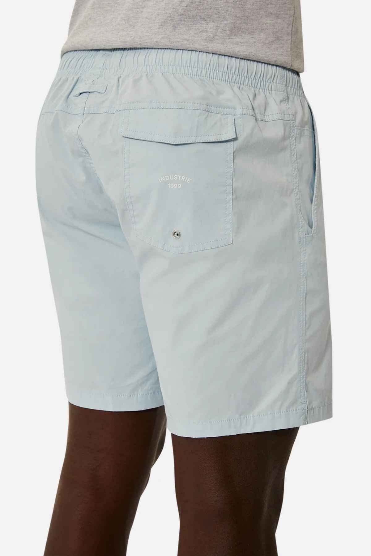 A person is sporting Industrie The Bahama Angler Short in light blue, featuring a drawcord waist and pocket details. The back pocket proudly displays a small label that reads "INDUSTRIE 1999." Paired effortlessly with a gray shirt, the ensemble stands out against the plain white background.