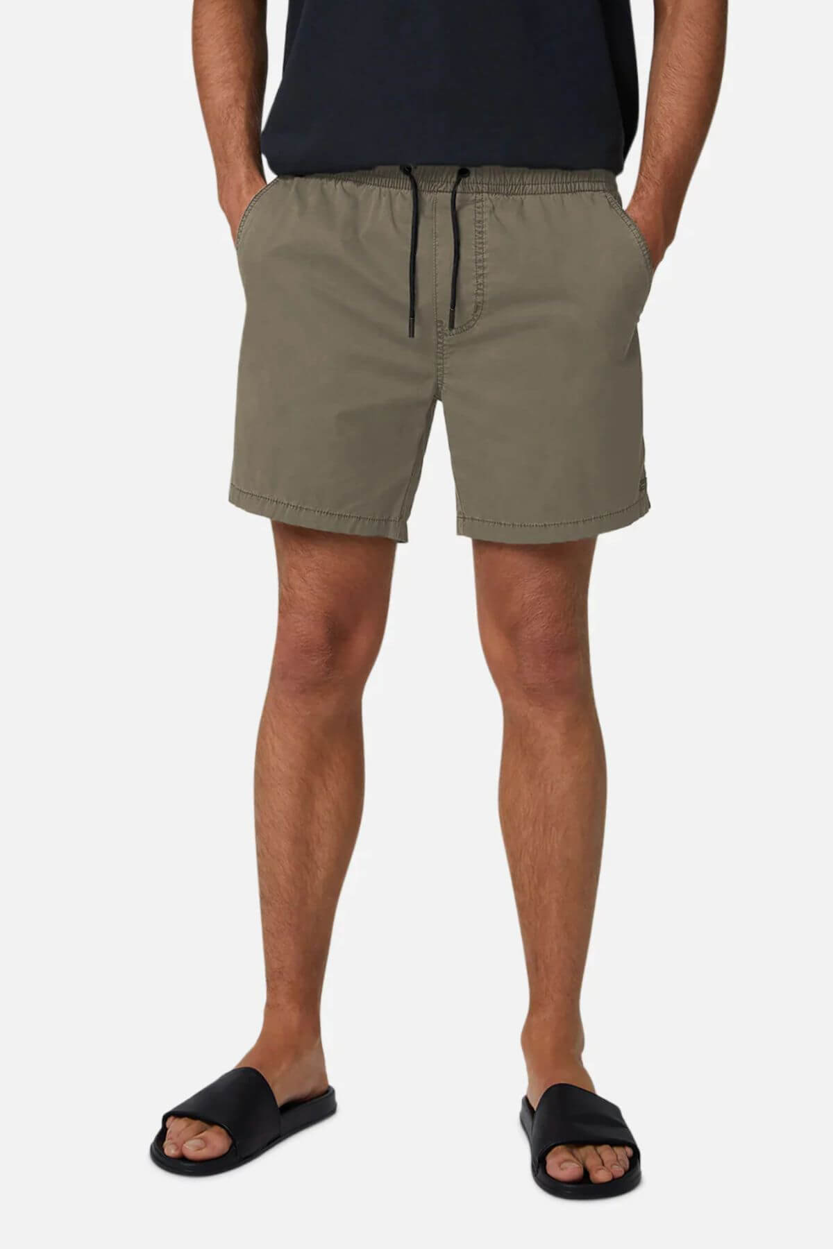 A person stands against a white background wearing Industrie The Bahama Angler Short in olive green, featuring an elastic waistband and drawstring, paired with a black T-shirt and black slide sandals. The shorts subtly display the Industrie logo.