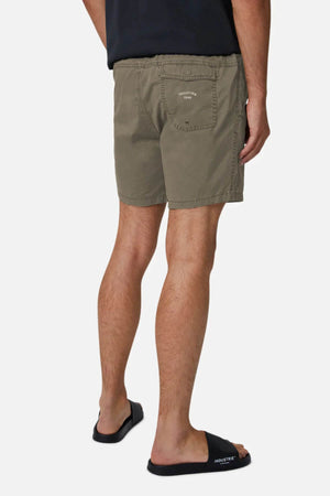 A person in olive green Industrie The Bahama Angler Shorts with an elastic waist and a subtle logo on the back pocket, paired with black slides and a partially visible black shirt, stands facing away against a plain white background.