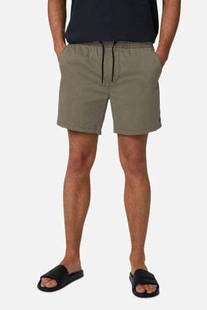 A person stands against a white background wearing Industrie The Bahama Angler Short in olive green, featuring an elastic waistband and drawstring, paired with a black T-shirt and black slide sandals. The shorts subtly display the Industrie logo.