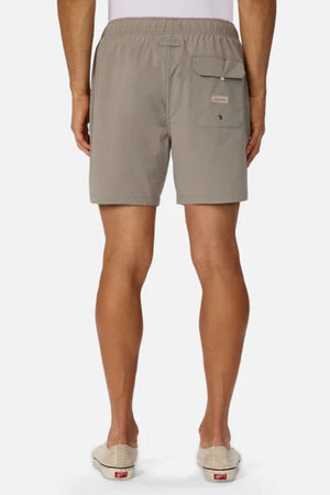 A person wearing Industrie The Barbados Swim Short in taupe, featuring an elastic waistband and a right back flap pocket, with light slip-on shoes, shown from the back against a plain background.