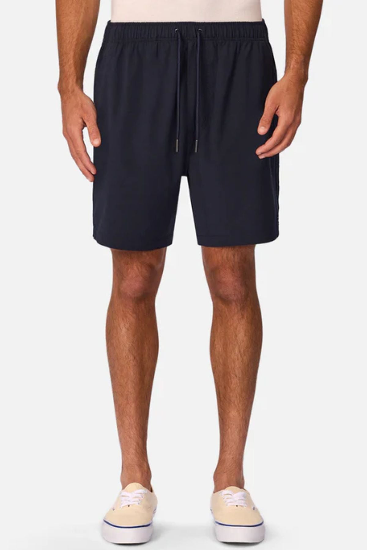 A person wears Industrie The Barbados Swim Short in navy blue, featuring an elastic waistband and drawstring. Beige slip-on shoes complete the look. Only the lower body is visible, with arms relaxed at their sides.
