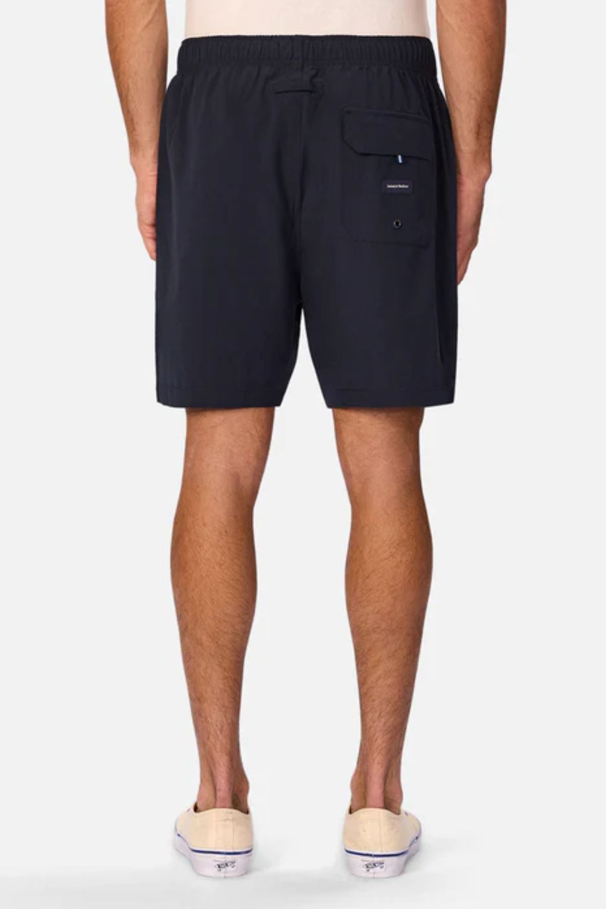 A person stands with their back to the camera, wearing Industrie The Barbados Swim Short—black, with an elastic waistband and back pocket—ideal men's beachwear. White slip-on shoes complete the relaxed look against a light background.