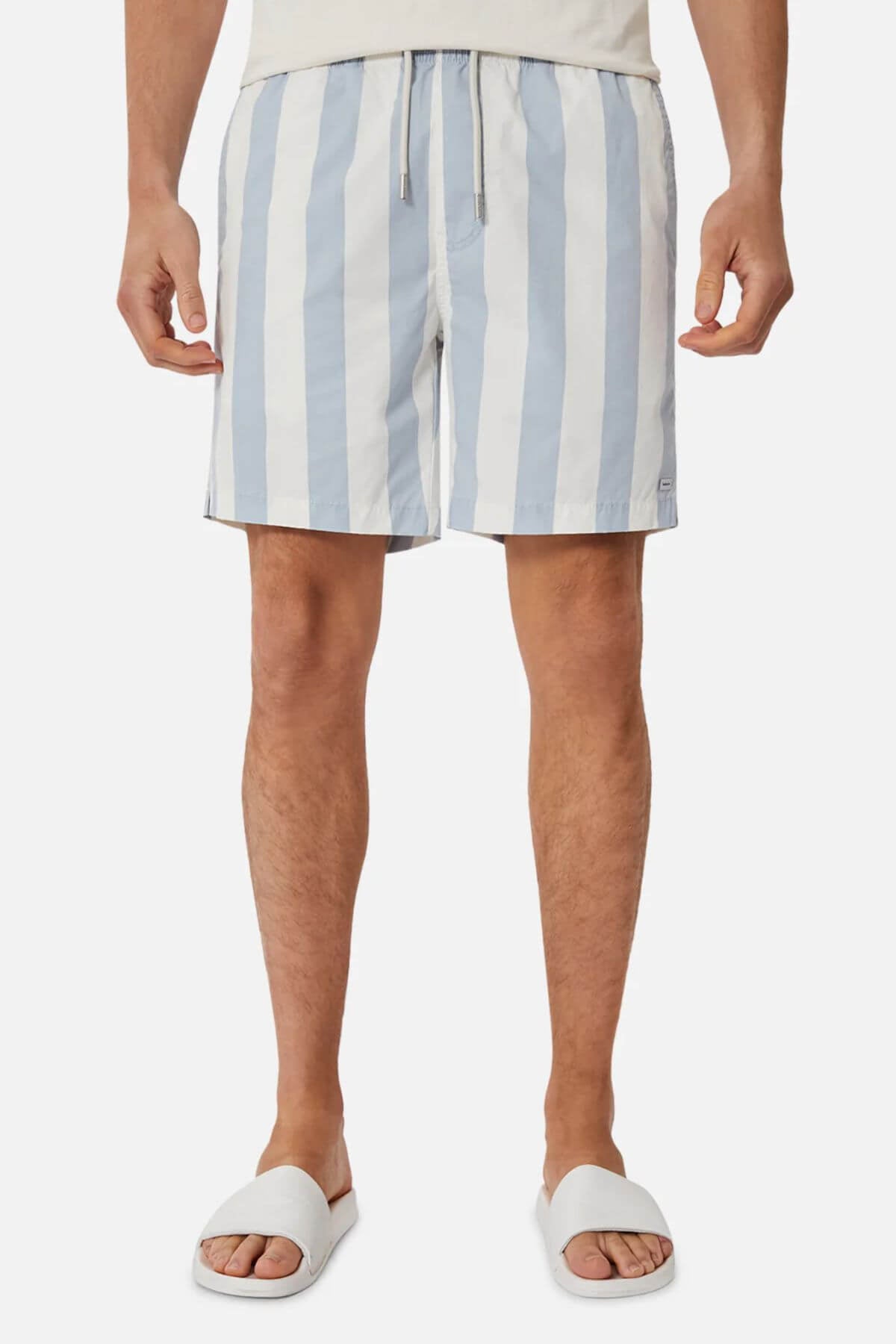 A person stands against a plain background wearing Industrie The Beverly Bahama Short, which features blue and white vertical stripes and an adjustable fit, paired with a light shirt and white slide sandals.
