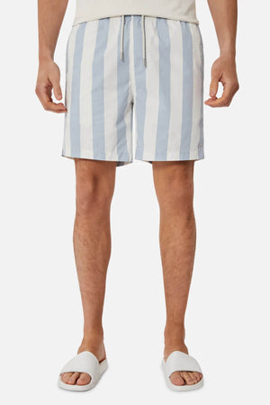 A person stands against a plain background wearing Industrie The Beverly Bahama Short, which features blue and white vertical stripes and an adjustable fit, paired with a light shirt and white slide sandals.