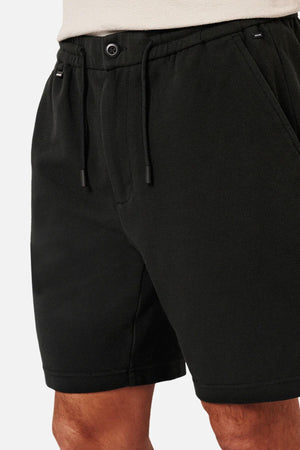 Close-up of a person wearing the Industrie The Broadway Short in dark green, featuring a relaxed fit with an elastic waistband, drawstring, button, side pockets, and crafted from stretch fabric for comfort. They're paired with a light-colored shirt.