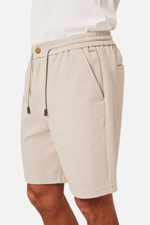 A person wearing Industrie The Broadway Short in light beige, featuring an elastic waistband, drawstrings, and side pockets for a relaxed fit. Paired with a white top; image is cropped to show the lower torso and upper legs.